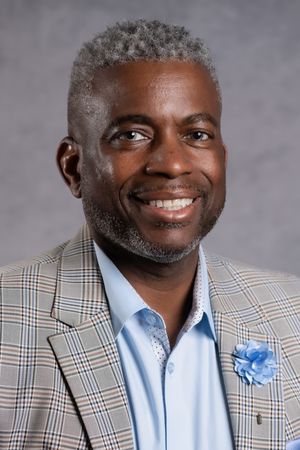 Smiling middle-aged man with gray hair wearing a plaid blazer and blue shirt with a floral lapel pin.