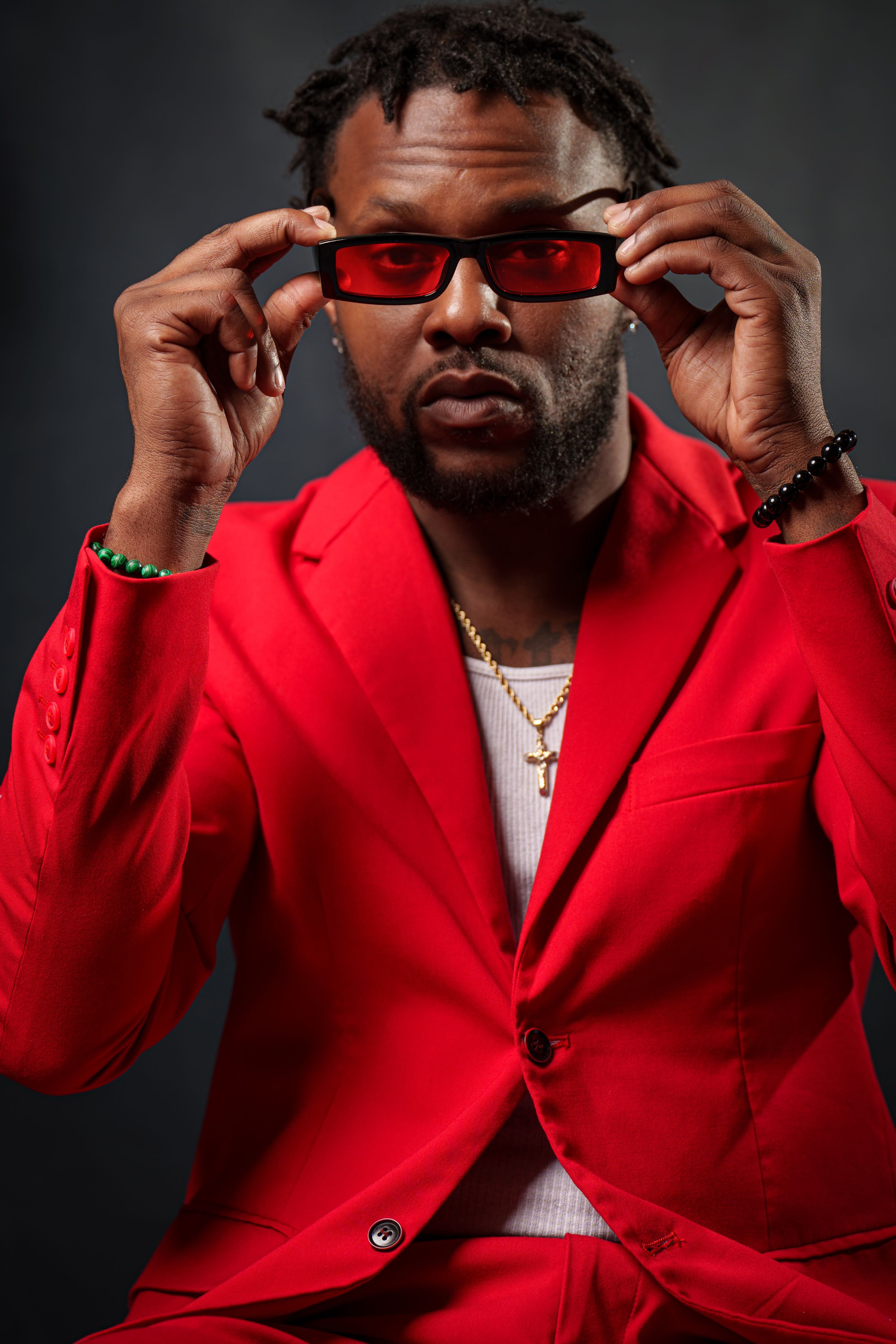 Man in red suit adjusting red-tinted sunglasses, wearing gold chain necklace and beaded bracelet, dark studio background.
