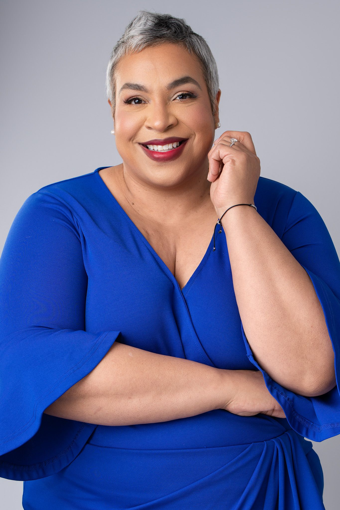 A smiling woman with short gray hair wearing a bright blue wrap dress poses confidently against a gray studio background.