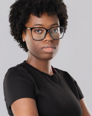 Young Black woman with natural hair and black rectangular glasses wearing a black t-shirt against a gray background.