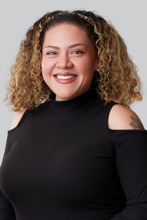 Smiling woman with curly blonde hair wearing a black cold-shoulder turtleneck top, with a tattoo on her shoulder.