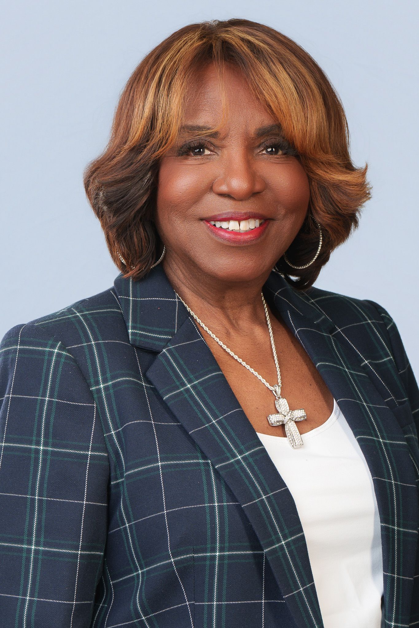 Professional woman in plaid blazer and cross necklace smiling against light blue background.