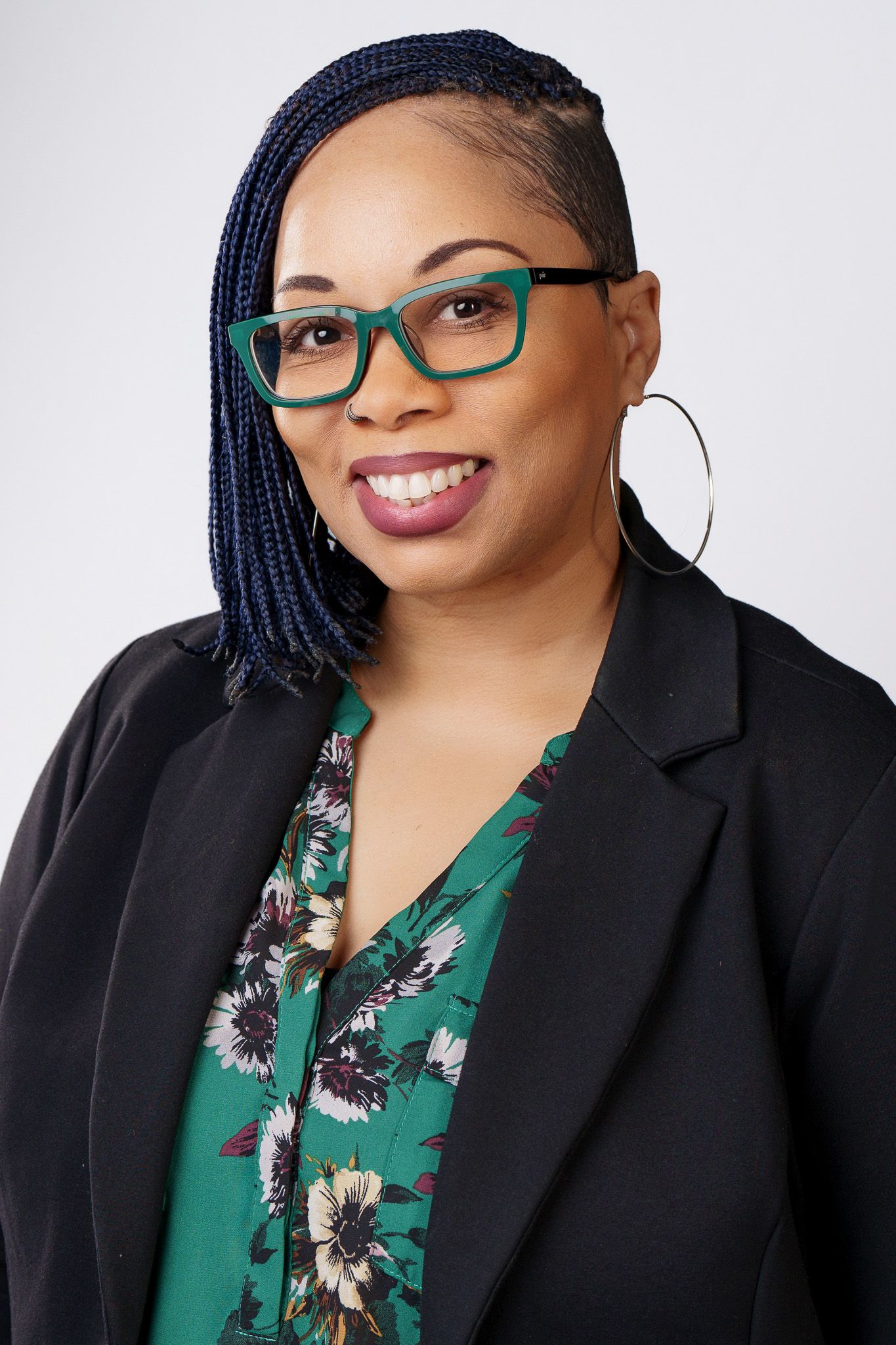 Professional woman with blue braids, green glasses, floral blouse, and black blazer smiling in a studio portrait.