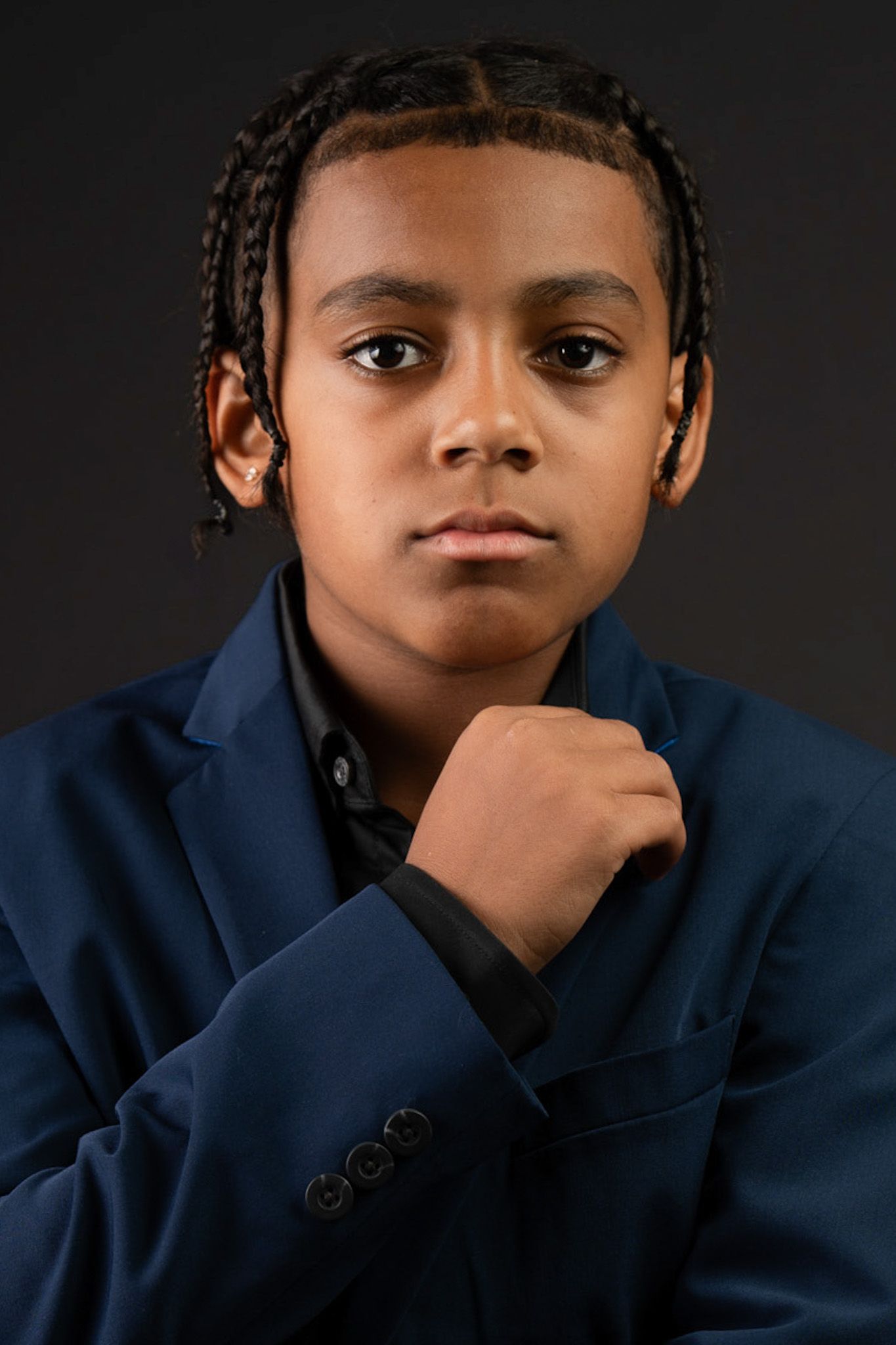 Young boy in navy blue blazer with braided hair poses confidently against dark background.