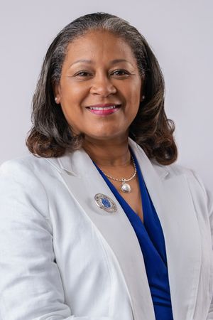 Professional headshot of a smiling woman in a white blazer and blue top, wearing a pearl necklace and brooch.