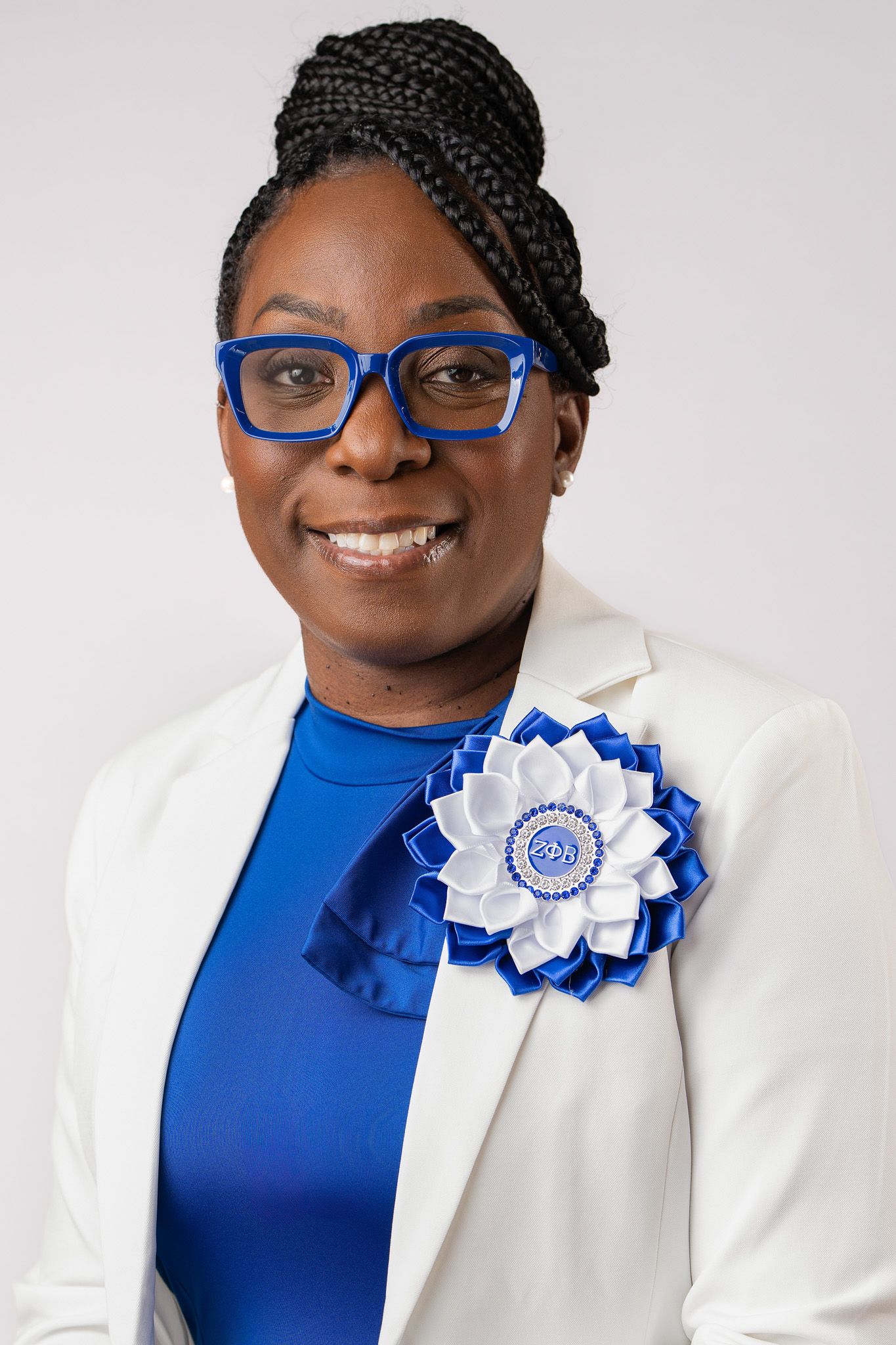 Professional woman in white blazer and blue top with decorative flower brooch, wearing blue glasses and braided updo hairstyle.