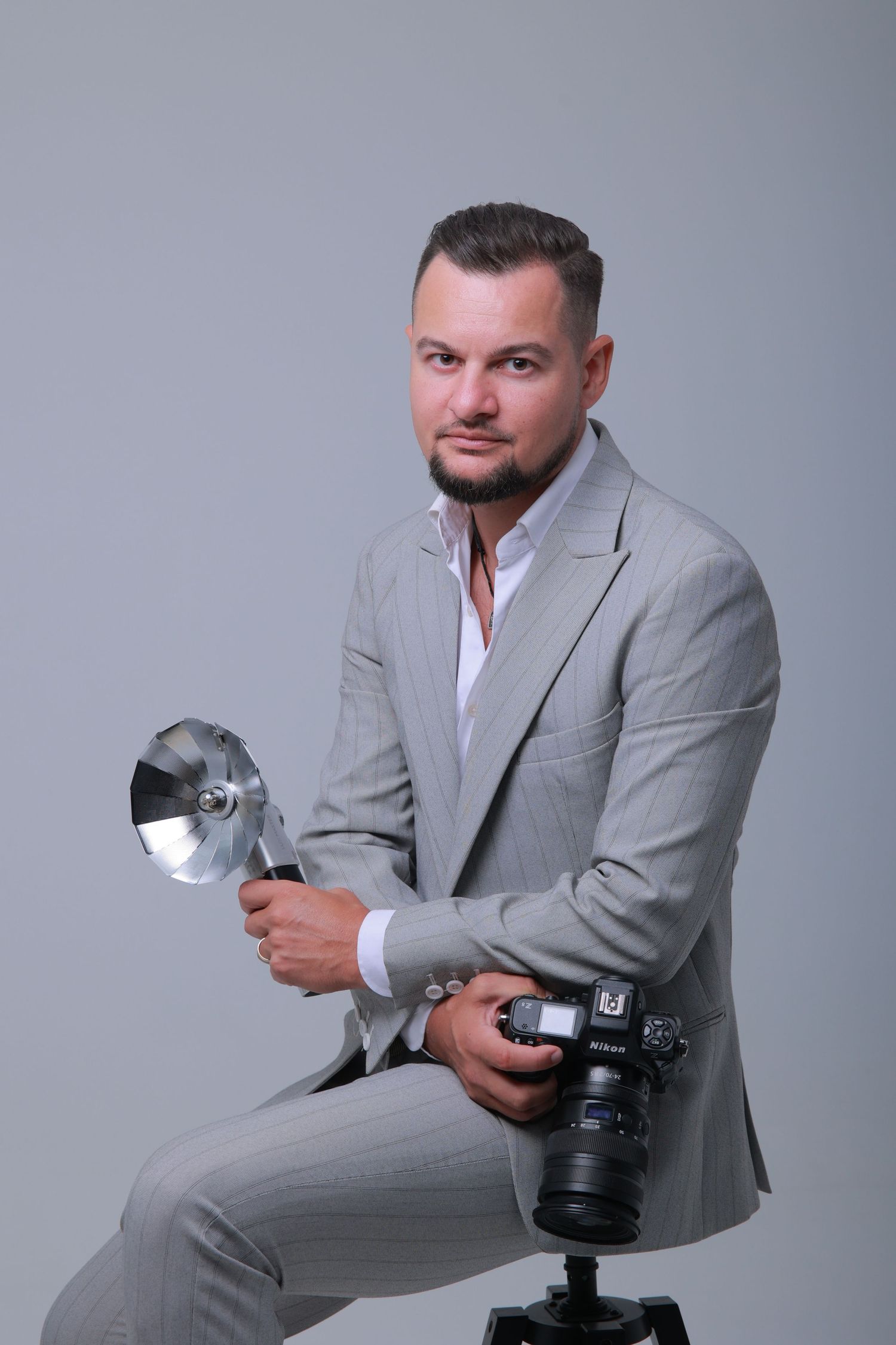 Professional photographer in formal attire holding camera equipment near curtained window.