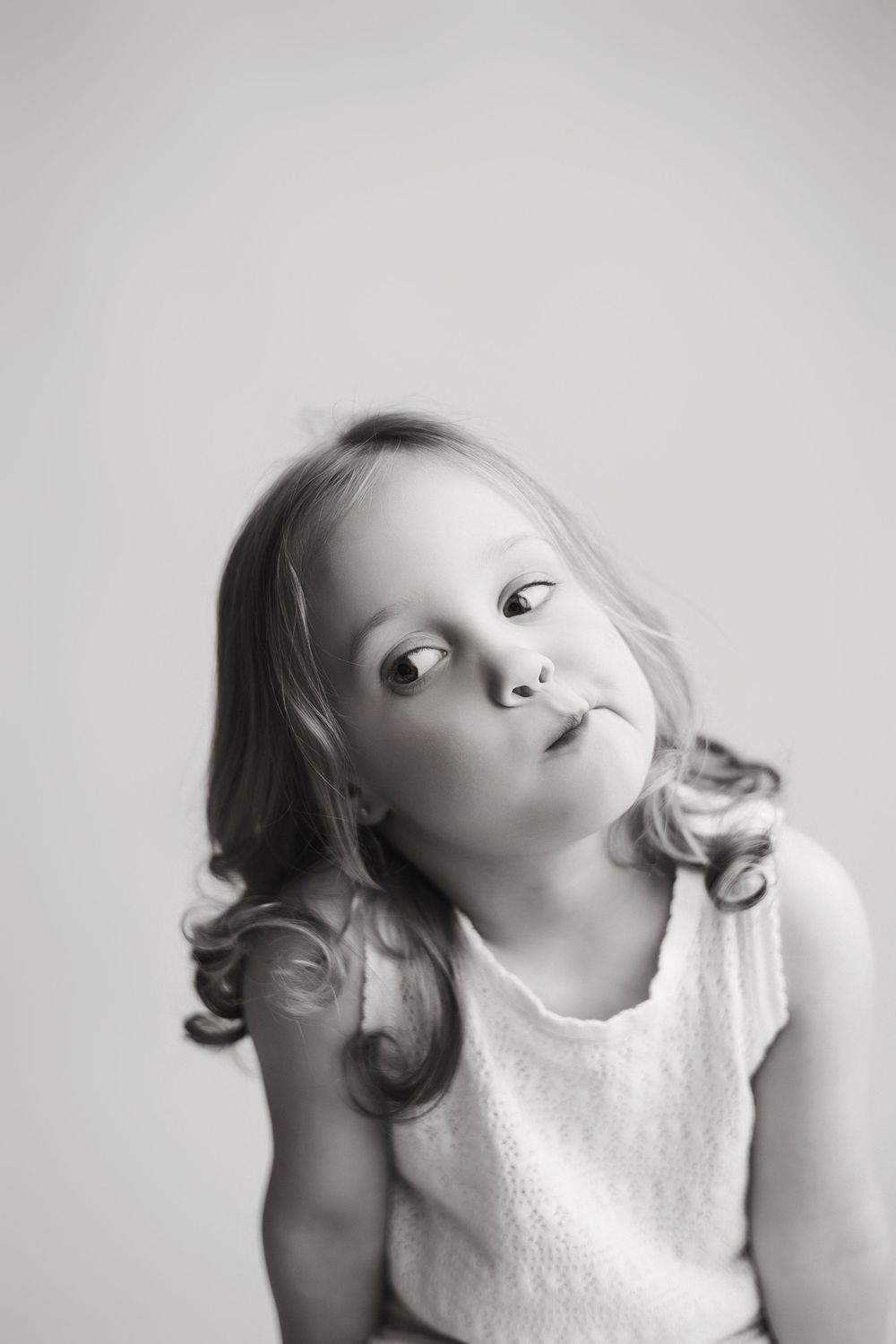Black and white portrait of a young child in a sleeveless white top looking up with curled hair.