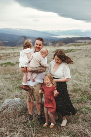 A loving family stands together on a hillside meadow with mountains and cloudy skies in the background.