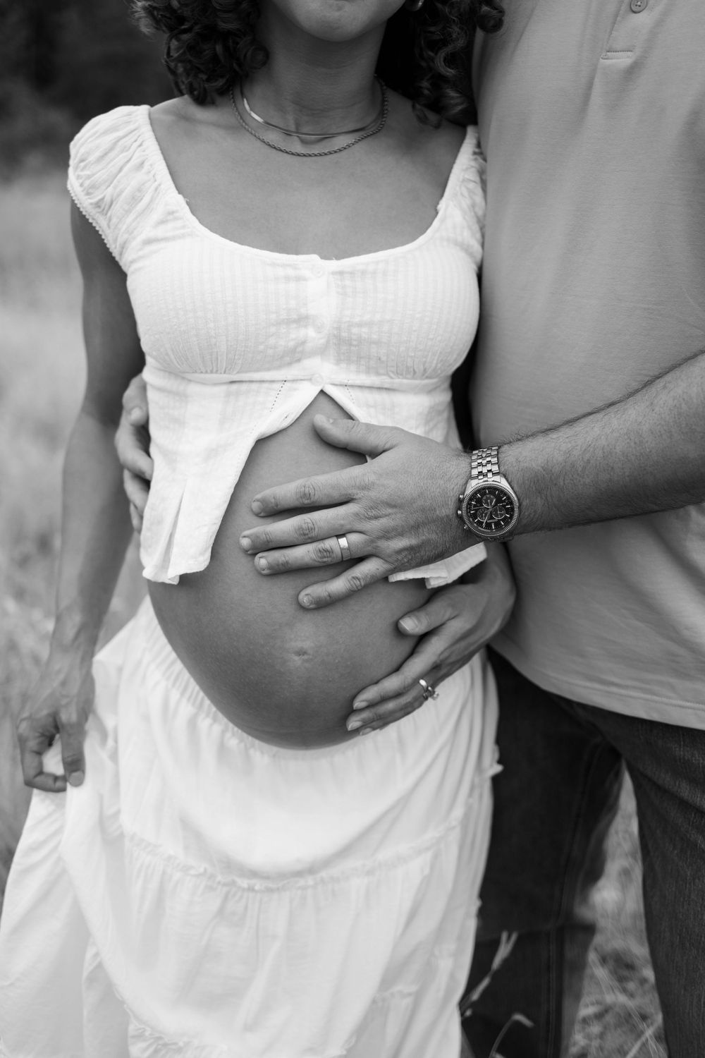 Black and white photo of pregnant woman in white dress with partner's hands cradling her baby bump outdoors.