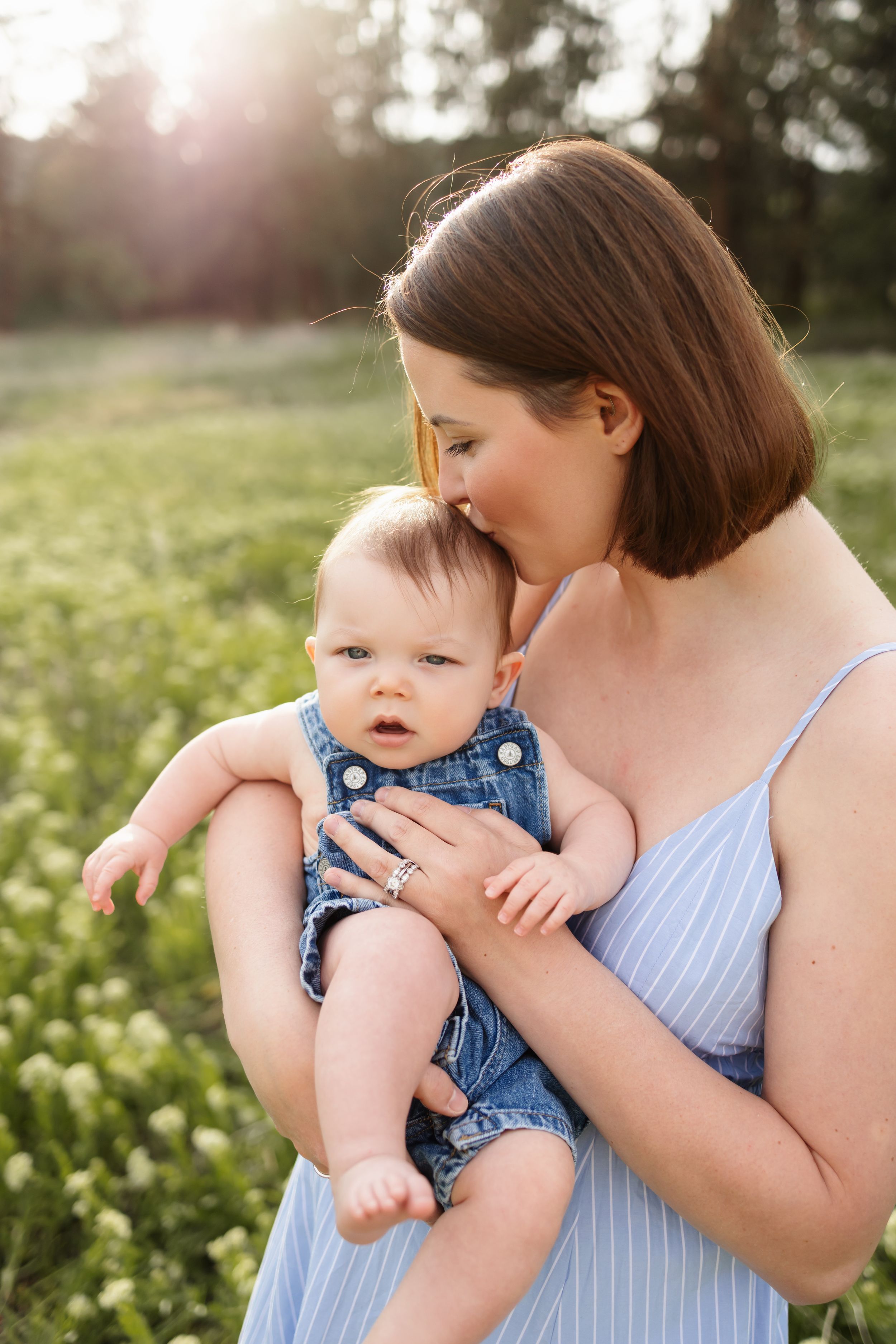 A mother in a blue dress kisses her baby in denim overalls outdoors in a sunlit green field.