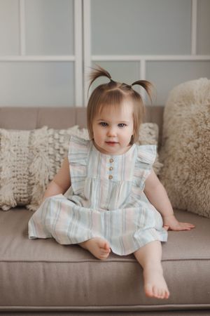 A happy toddler in a striped ruffled dress sits on a gray couch with decorative throw pillows.