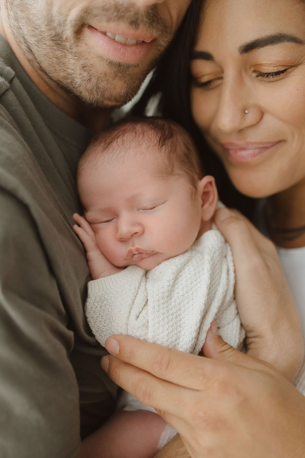 Parents lovingly cradle their sleeping newborn baby wrapped in a white knit blanket.