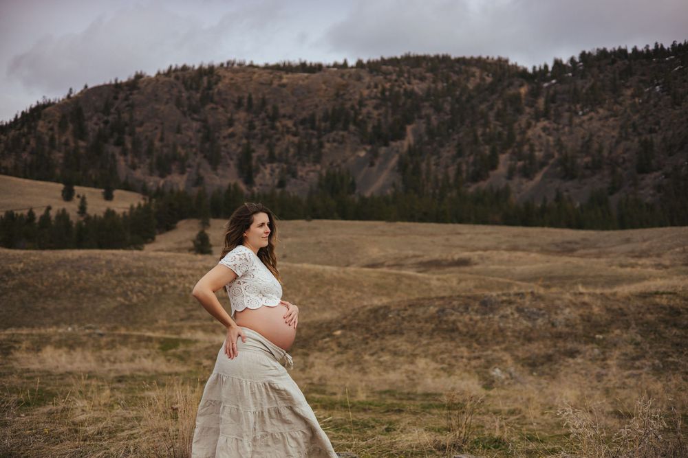 An expecting mother in a flowing white dress stands in a scenic mountain meadow at sunset.