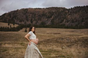 An expecting mother in a flowing white dress stands in a scenic mountain meadow at sunset.