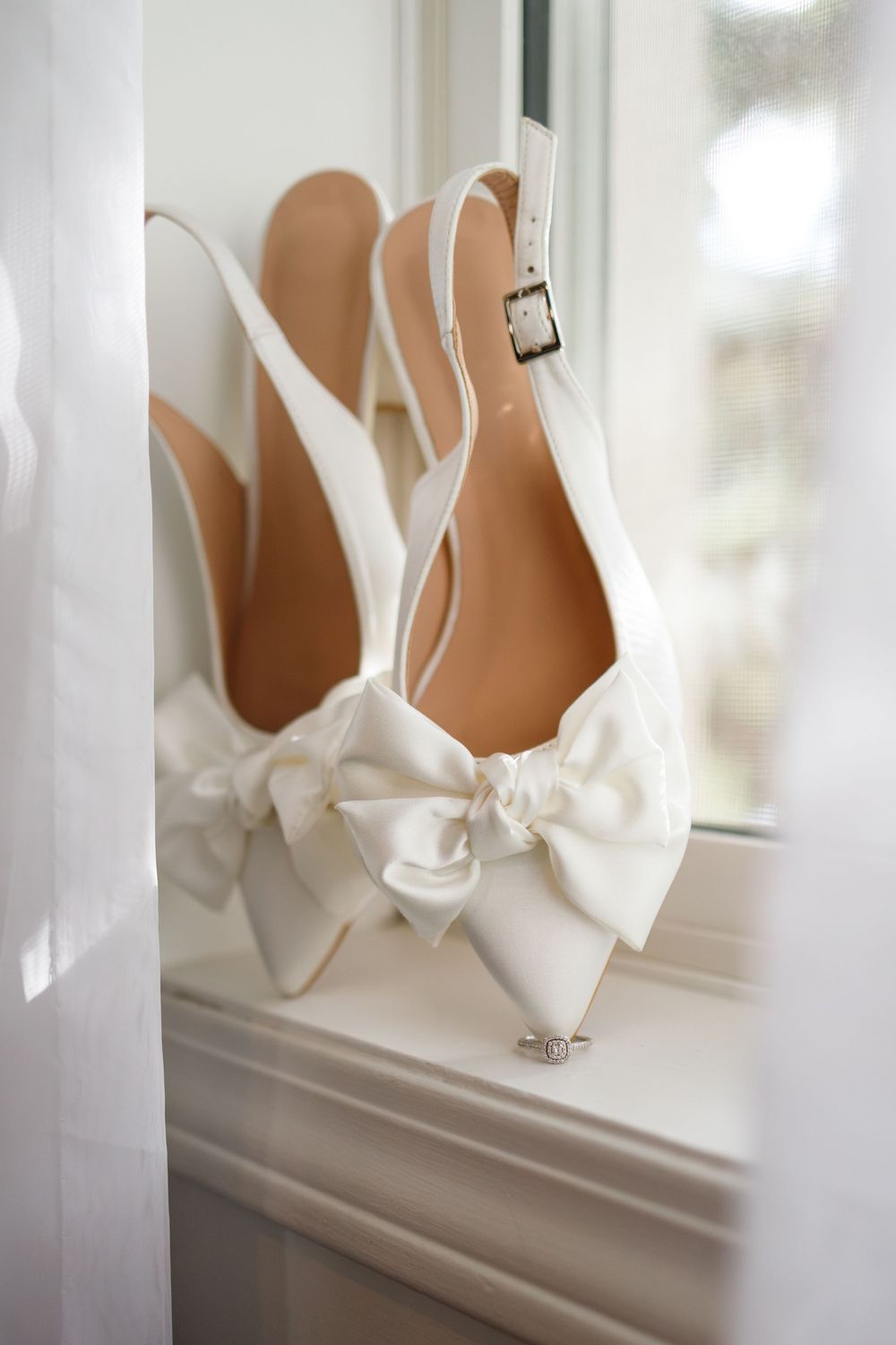 White satin bridal heels with elegant bow detail and ankle strap displayed on window sill.