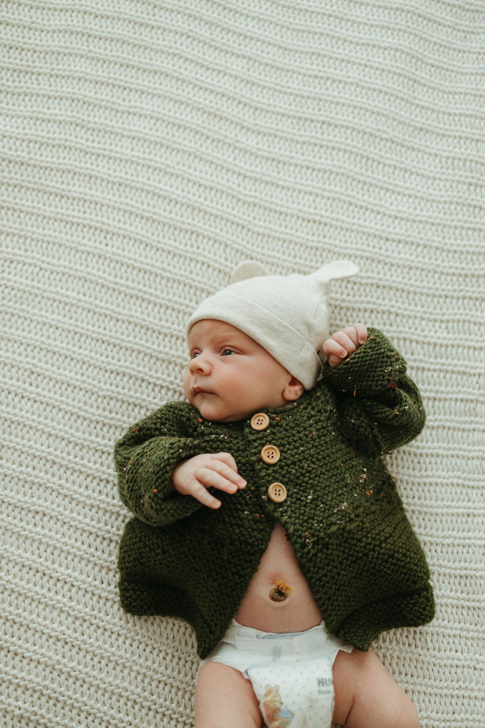 Newborn baby in green knit cardigan and white knotted hat lying on cream textured blanket.