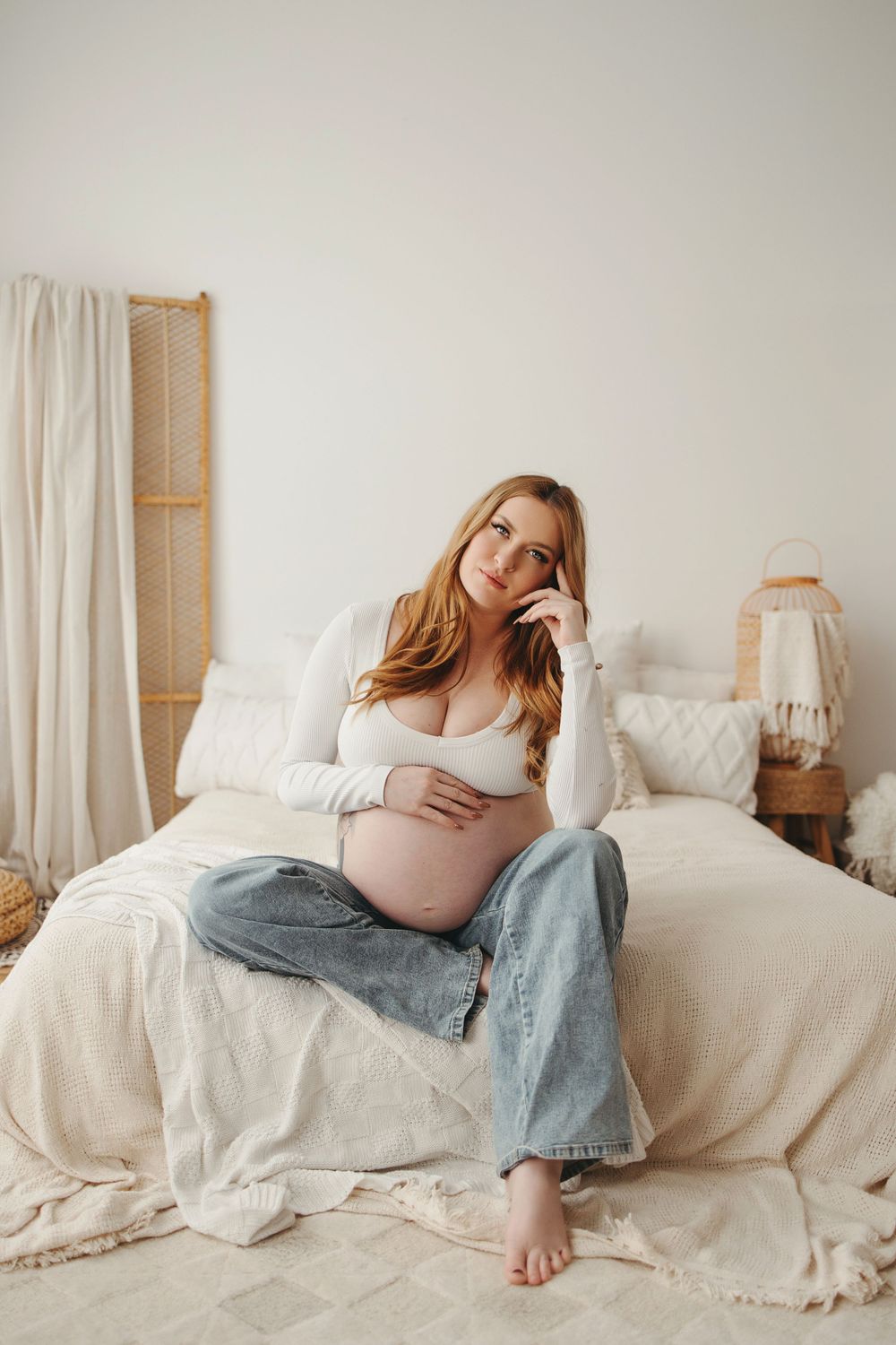 Expectant woman in jeans and white top relaxes on cozy white bed with neutral decor and natural lighting.