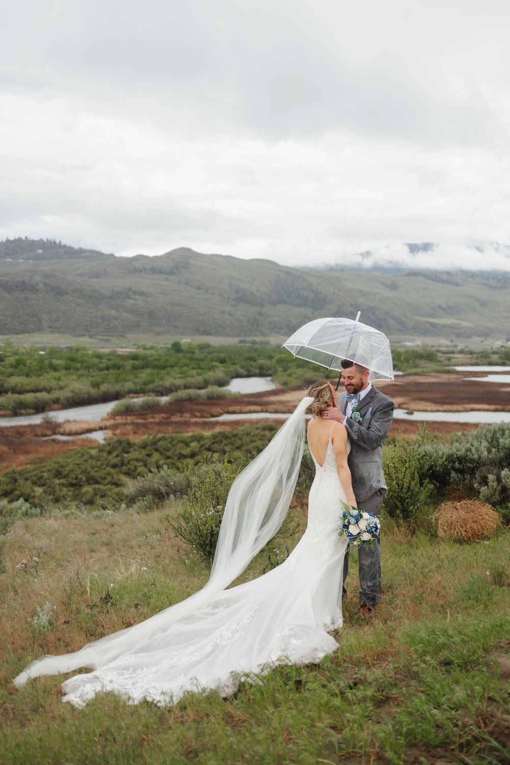 A couple shares an umbrella on their wedding day amid rolling hills and overcast skies with a flowing white dress train.