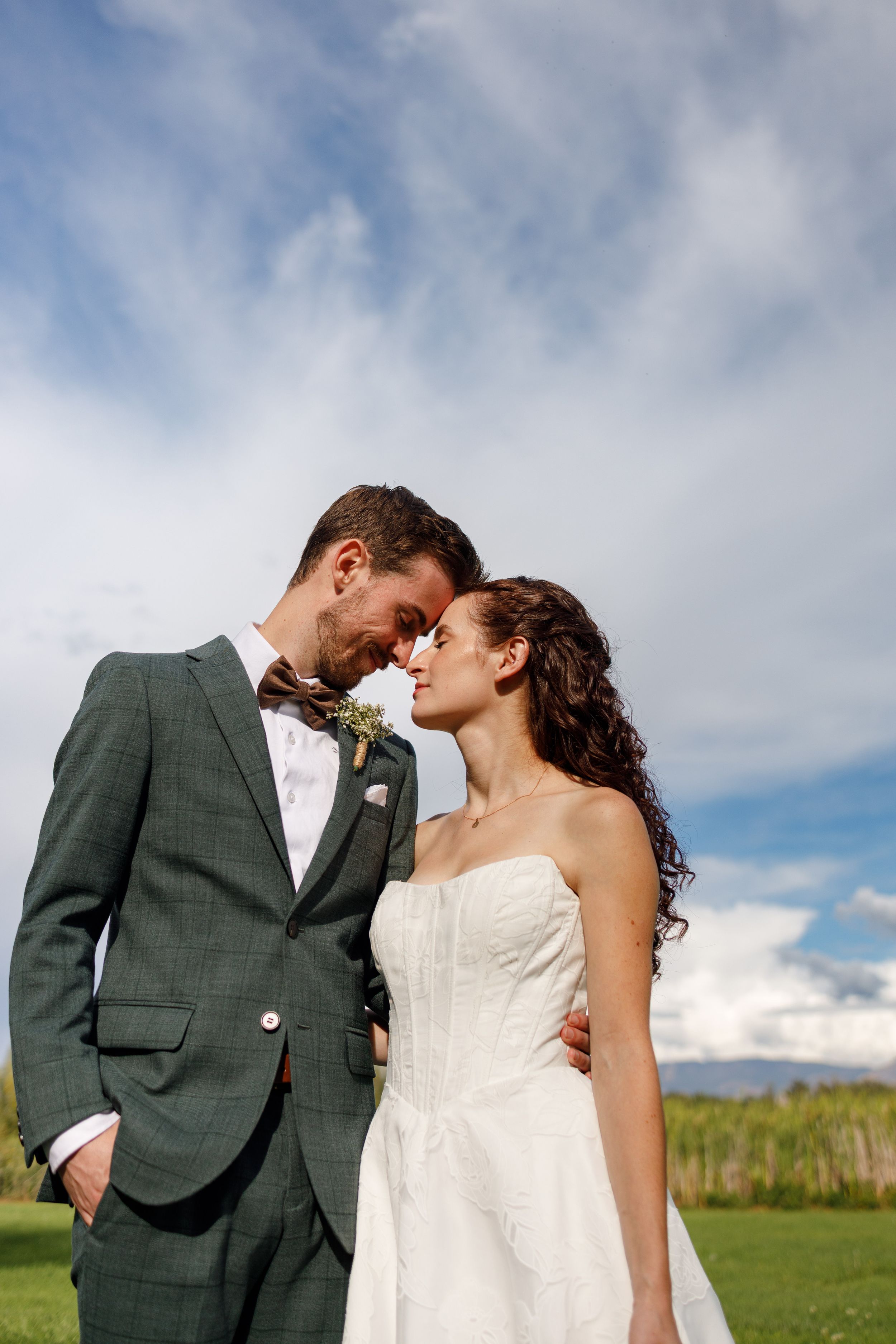 Bride and groom in green plaid suit and white strapless gown touch foreheads romantically outdoors under a blue sky.