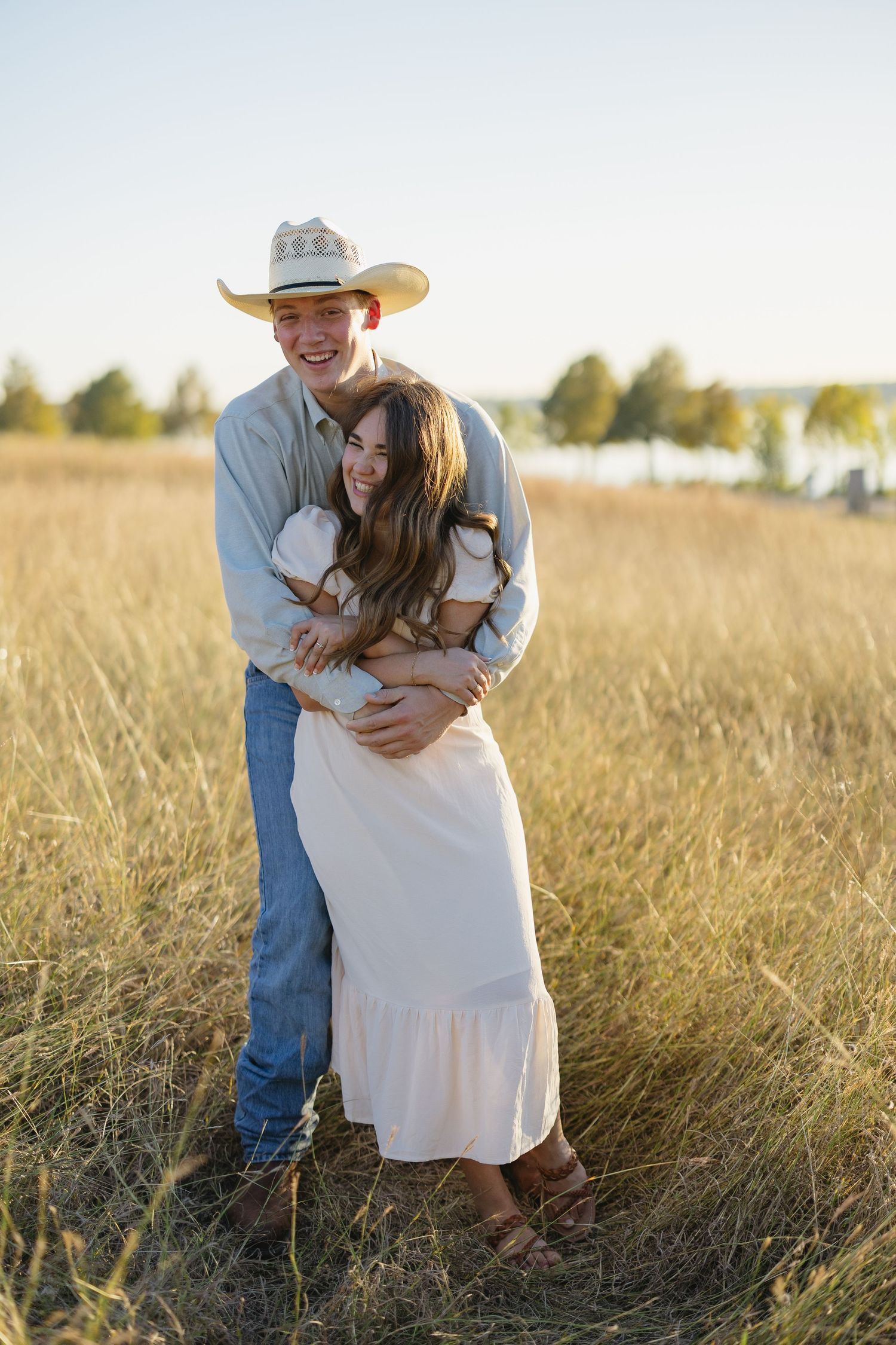 Happy couple embracing in golden field at sunset, man wearing cowboy hat and woman in white dress.