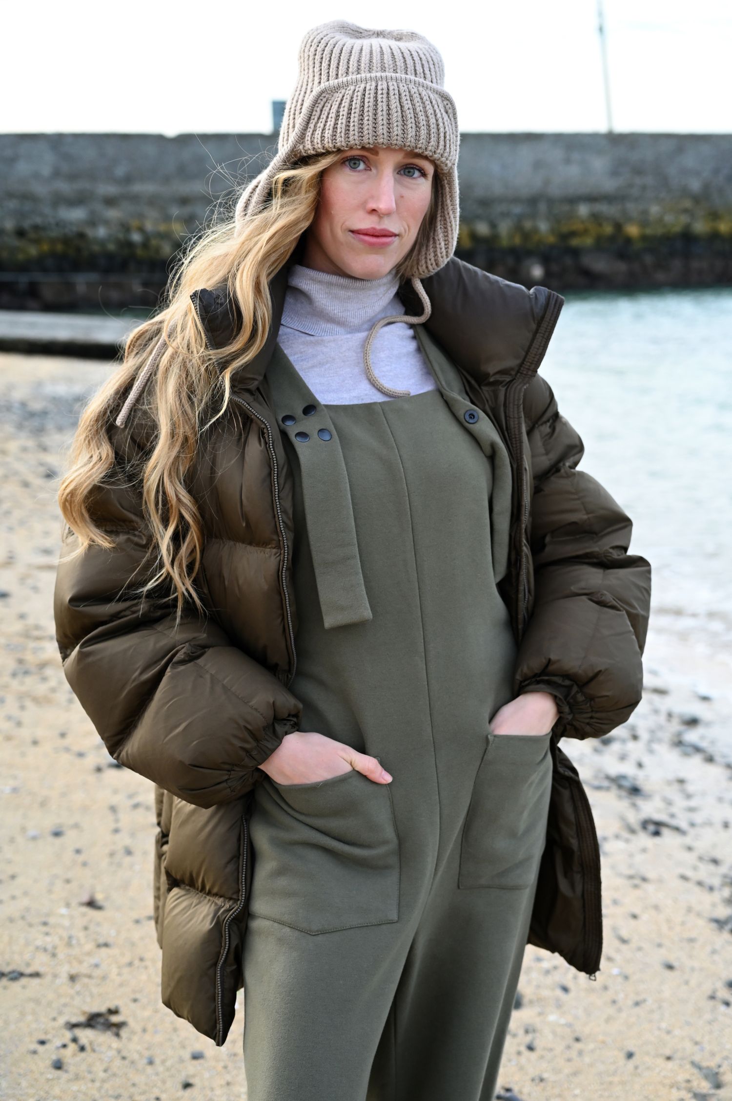 Woman wearing winter coat and knit beanie poses on rocky beach.