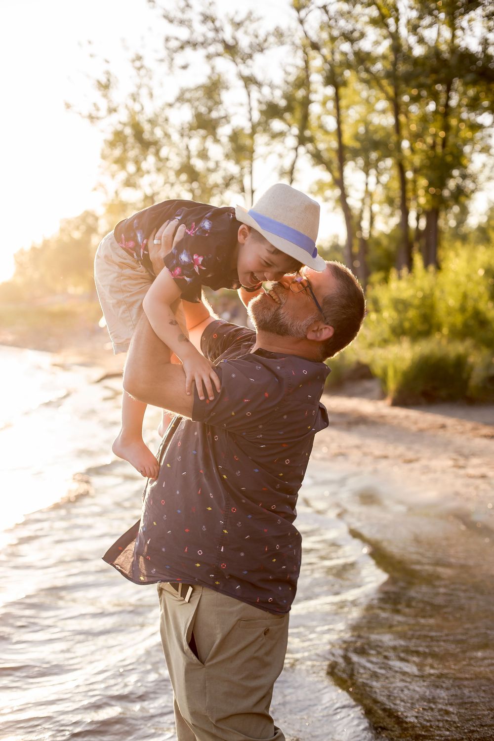 Father lifting and kissing young child wearing a hat on a sunny beach with trees in the background.