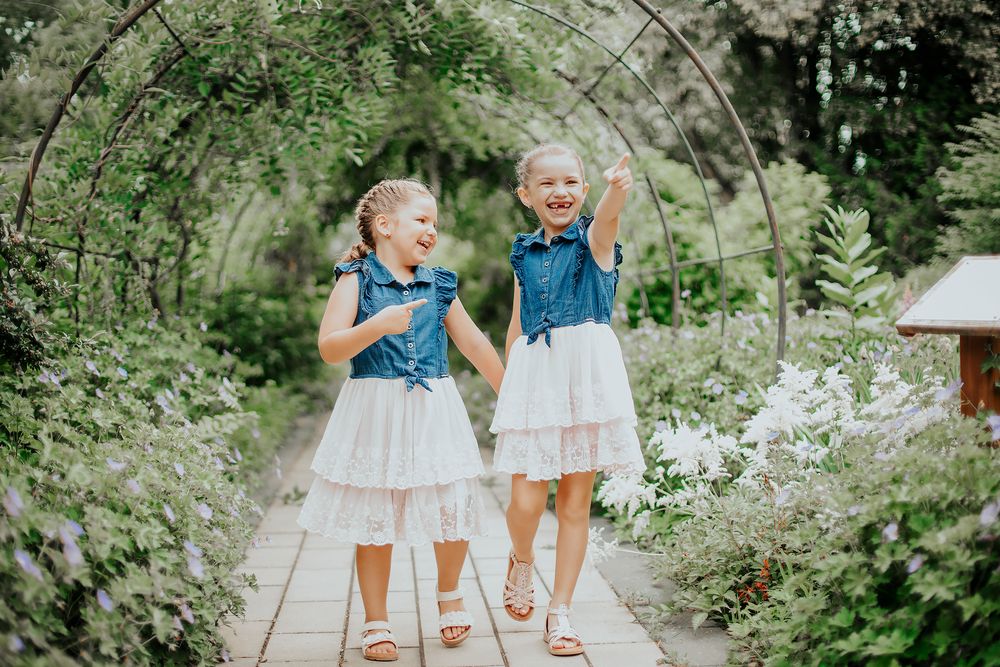 Two young girls in matching denim and white tulle dresses laugh and play under a garden archway.