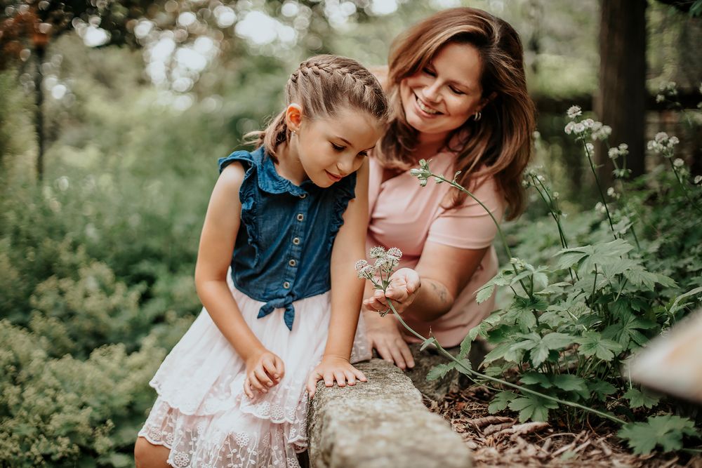 A mother and young girl explore nature together, examining plants near a log in a lush green forest setting.