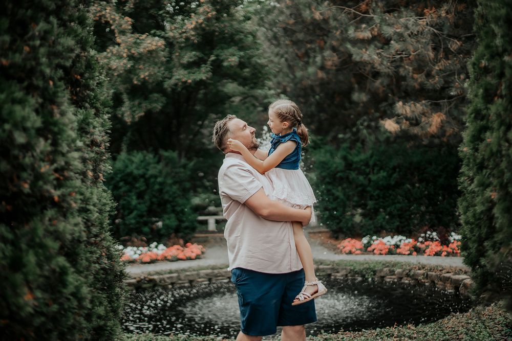 A father lovingly lifts his young daughter in a lush garden with a fountain and colorful flowers.