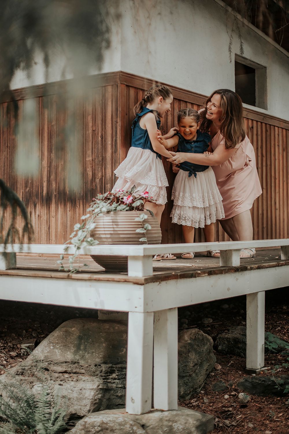 A mother lovingly interacts with her two young daughters in matching dresses on a wooden deck outside a rustic home.