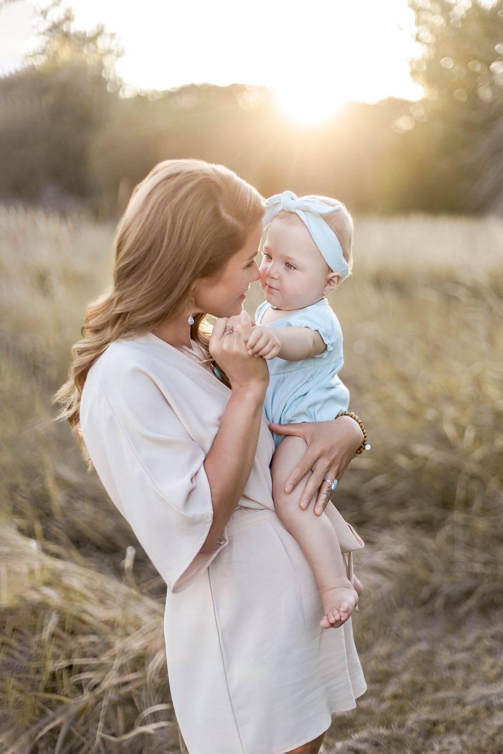 A tender moment between parent and baby captured in golden sunlight among tall grass at sunset.