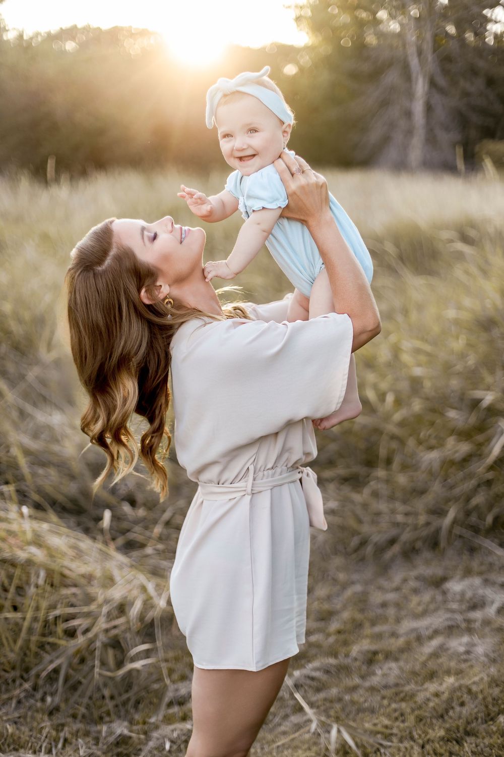 A parent in a beige sweater lifts and plays with a baby wearing blue in a sunlit outdoor field at golden hour.
