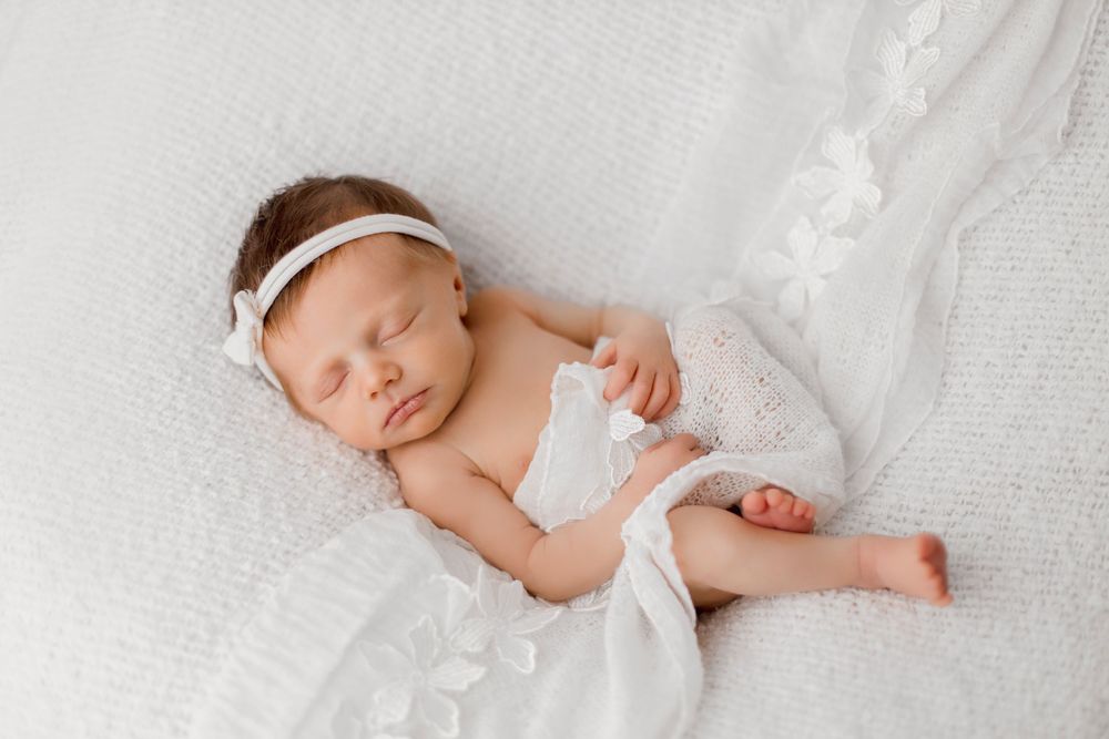 Peaceful newborn baby girl sleeping in white wrap and headband on soft white blanket in professional photo.
