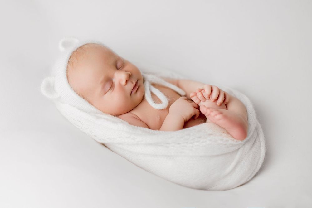 Sleeping newborn baby wrapped in white knit blanket wearing bear ear hat on white background.
