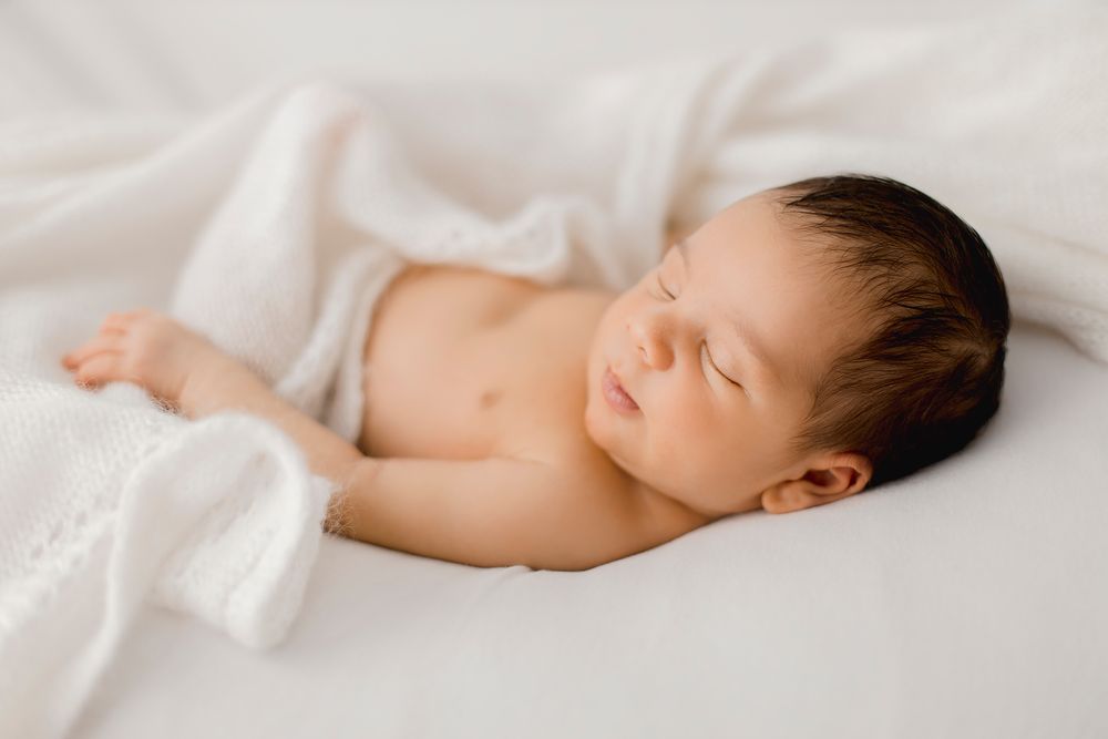 A peaceful newborn baby with dark hair sleeping on a soft white blanket in a serene newborn photography session.