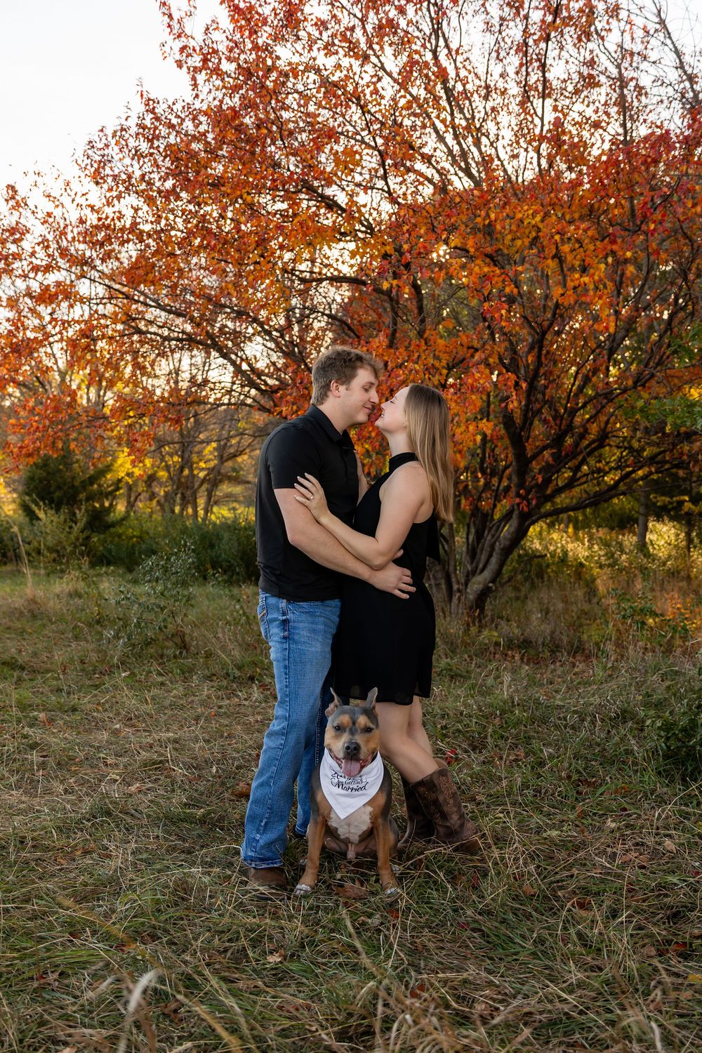 A couple embraces in an autumn field with their dog wearing a bandana.