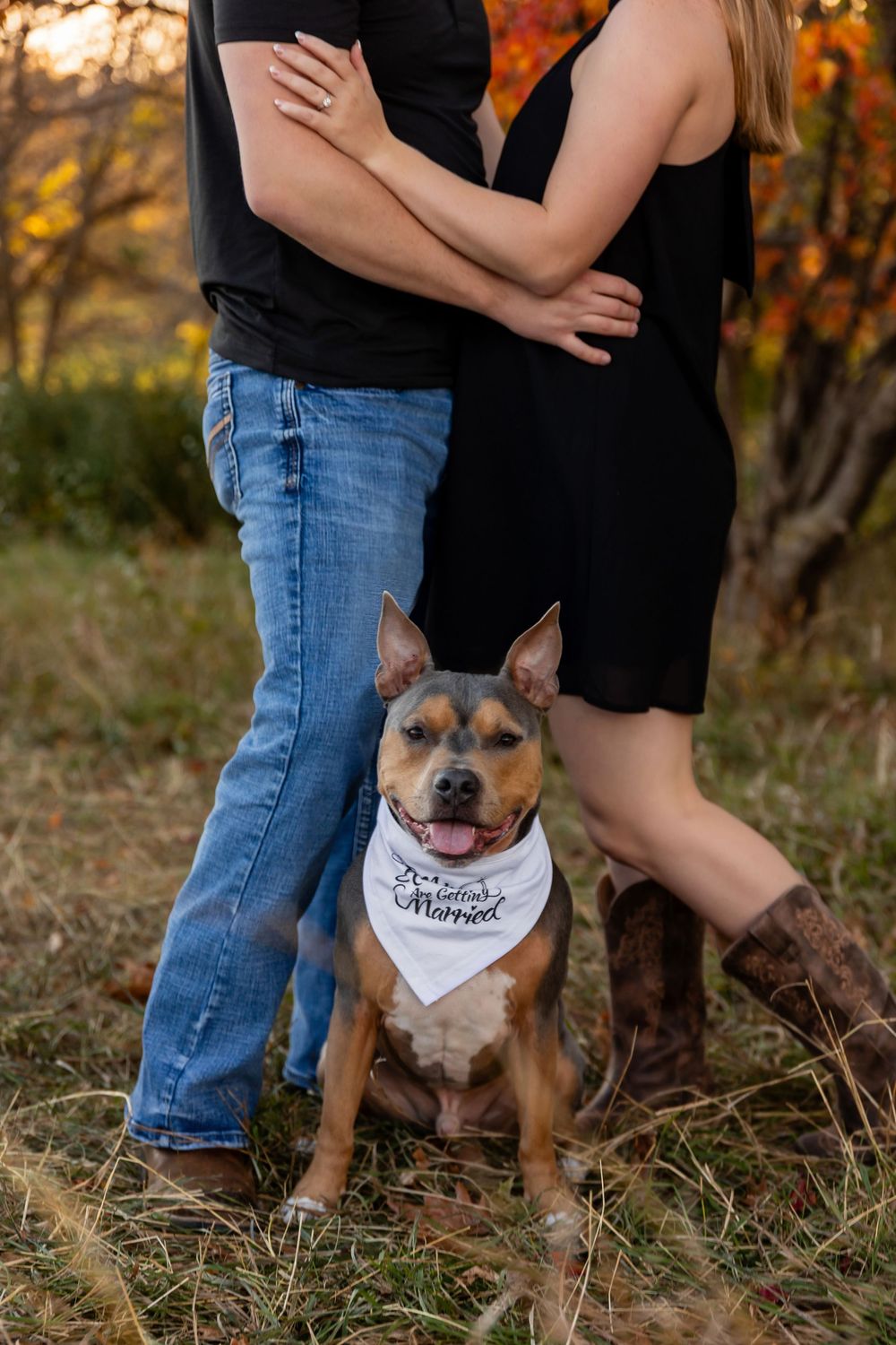 A couple poses in casual attire with their dog wearing a white bandana against fall foliage.