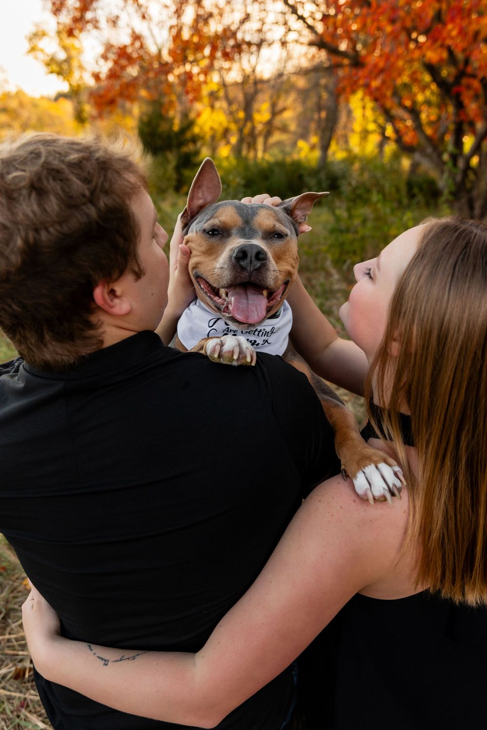 A couple holds their smiling dog between them during an outdoor autumn photoshoot.