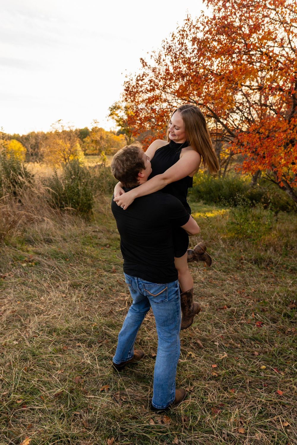 A couple embraces in a playful lift pose surrounded by autumn trees at sunset.