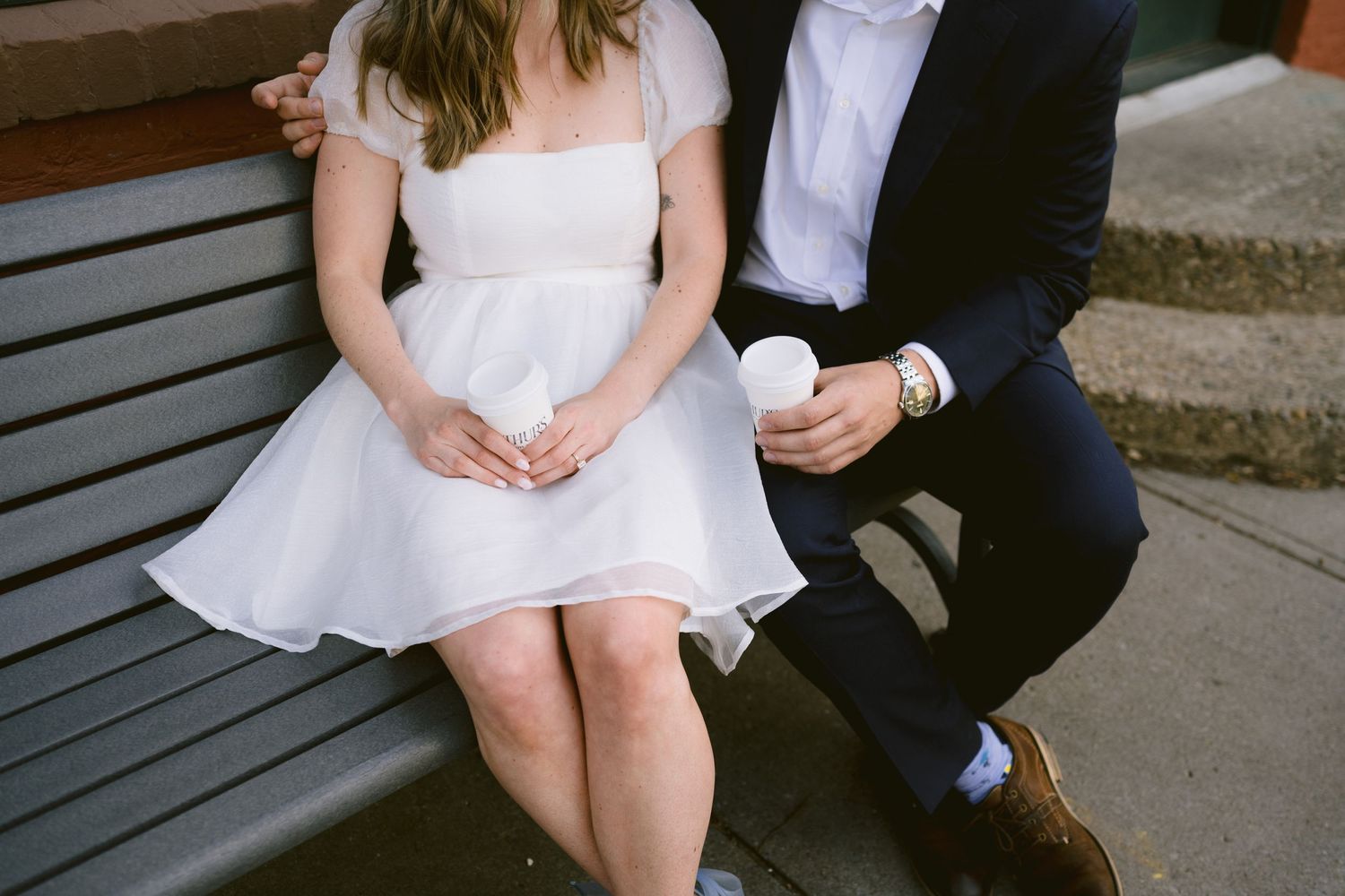 Couple in wedding attire sits closely on a bench, bride in short white dress holding a coffee cup.