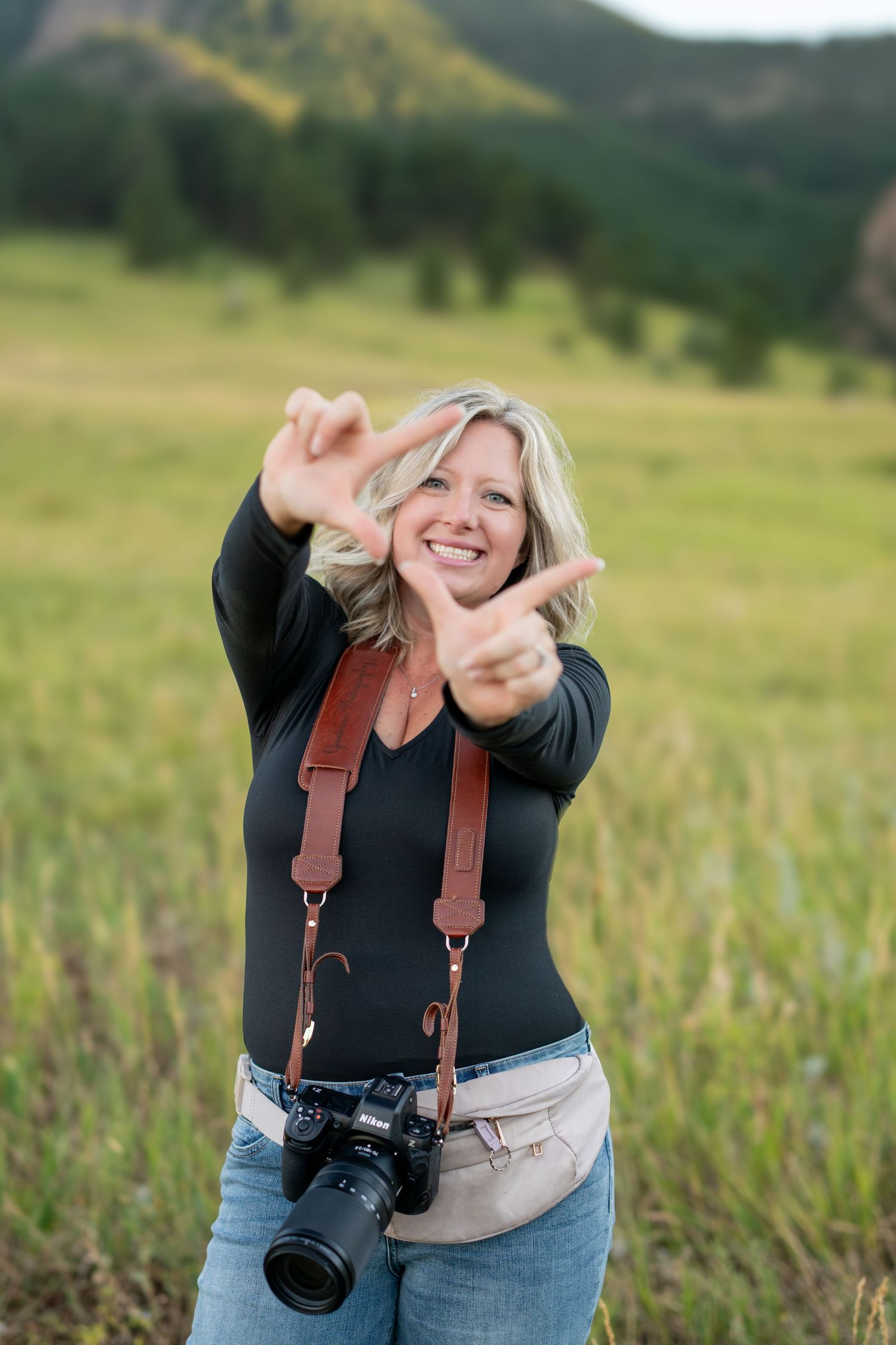Photographer with camera making peace sign gesture in mountain meadow.
