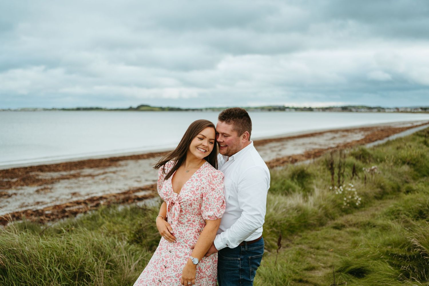 Couple embracing in coastal setting with grass and ocean in background.