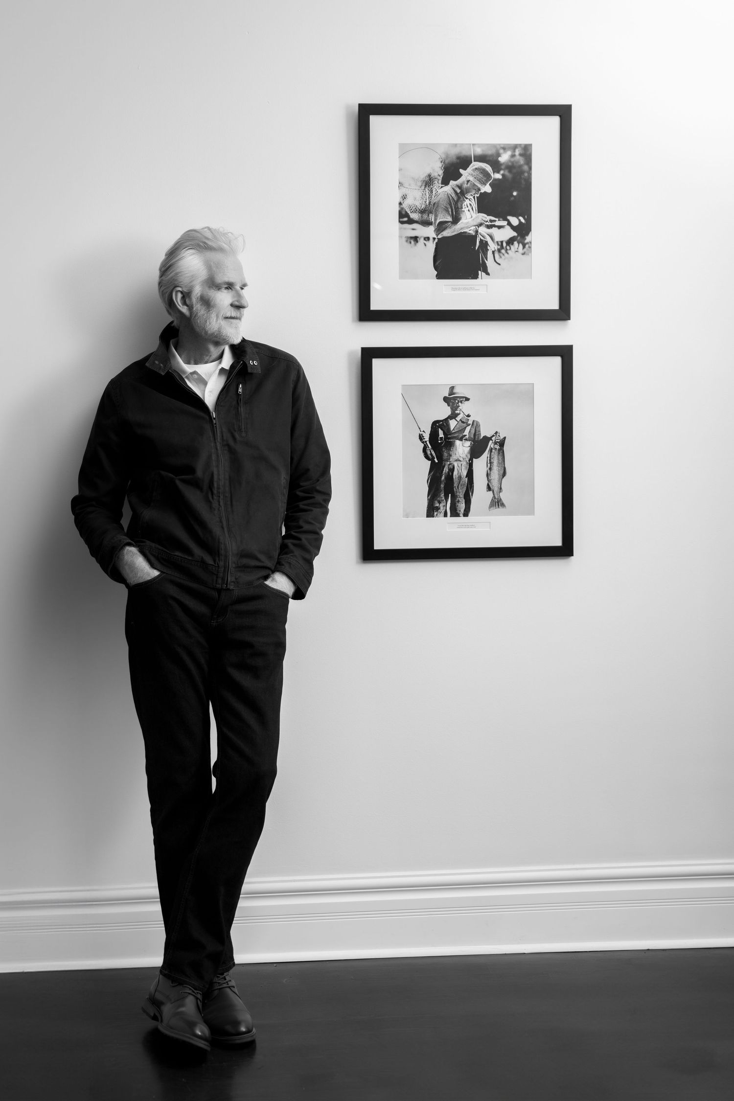 Black and white portrait of person standing against wall with framed artwork.