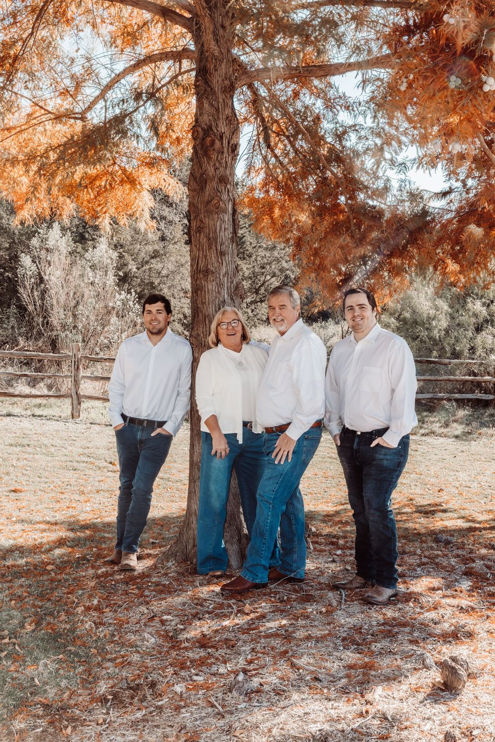Four people in white shirts and jeans pose together under autumn trees with orange foliage during an outdoor photo session.