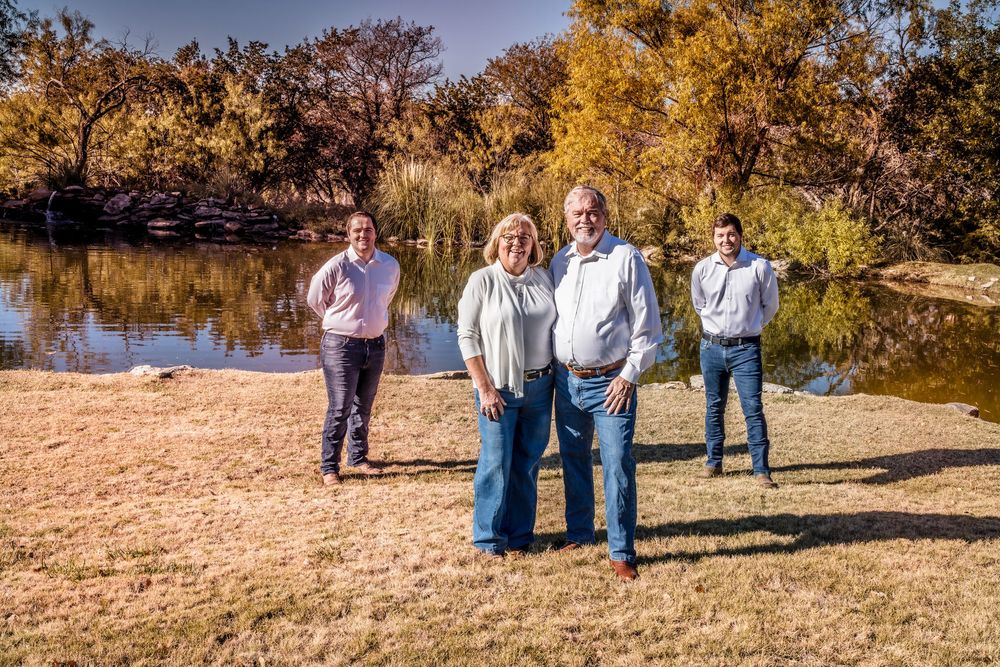 Group stands together on sandy shoreline beside lake with golden autumn trees in background on sunny day.
