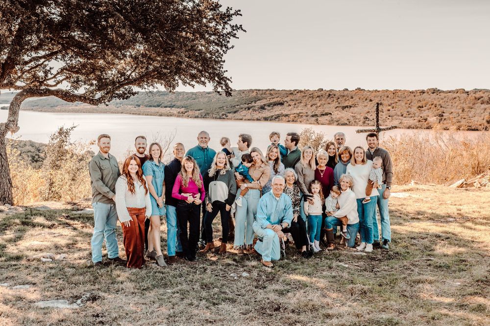 Large family group gathers outdoors for portrait under a tree in rustic countryside setting.