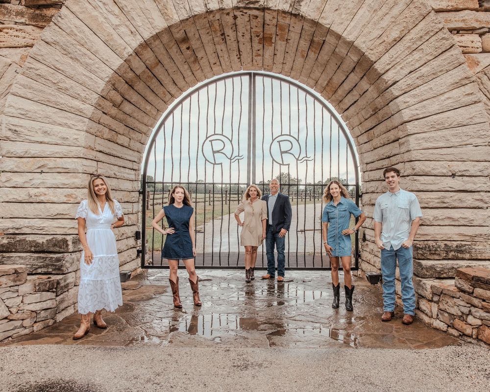 Group of people standing in front of a stone archway with decorative metal gate during a rainy day.