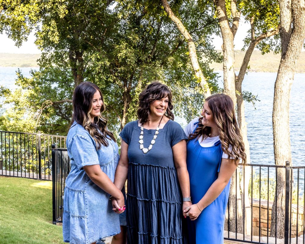 Three people in coordinating blue dresses share a lighthearted moment on a grassy lawn by a lakeside fence.