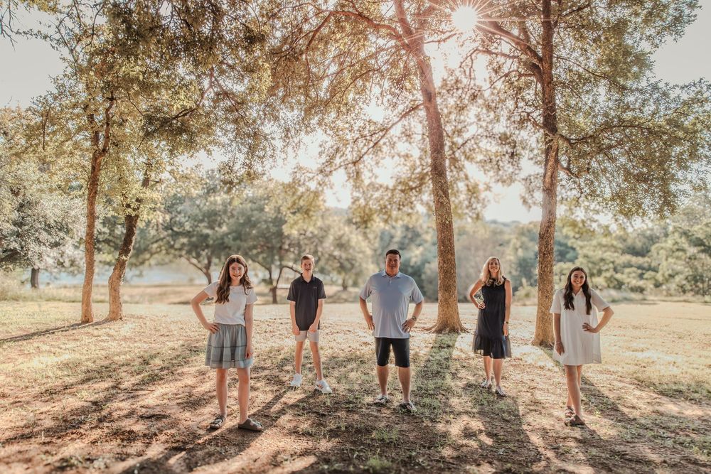 A group of people stand together under tall trees in a sunlit park during golden hour.